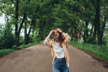 A stylish girl, a woman stands in a tunnel of green trees. Warm summer day. The wind blows the girl's hair and clothes. The girl is happy