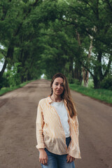 A stylish girl, a woman stands in a tunnel of green trees. Warm summer day. The wind blows the girl's hair and clothes. Girl looking at the camera
