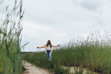 A girl, a woman walks towards the camera along a road in the middle of a field of green wheat. He dreams with his eyes closed and his arms spread out to the sides like wings.