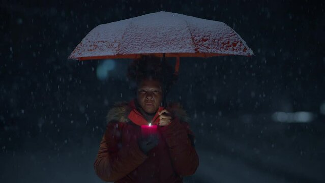 Young African Woman Standing on Street at Night During Snow Fall with Umbrella