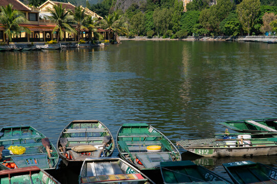Dozens of tin row boats lined up tied together on shore in Tam Coc in the Ninh Binh region of Vietnam. The town of Tam Coc in the background