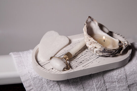 Guash massager and roll for facial massage lie on a light stand with a candle in the oyster sink, which stands on the bath cloth in the bathroom
