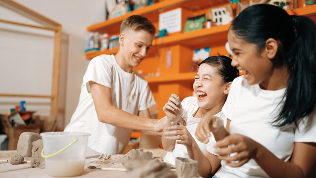 Handsome student playing with friend while put the clay on shirt at class. Group of diverse children working or modeling vase. Happy boy put water to young girl while laugh with happy. Edification.