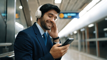Obraz na płótnie Canvas Smart business man listening music by headphone while waiting train at train station with blurring background. Skilled project manager enjoy listen relax sound while holding mobile phone. Exultant.