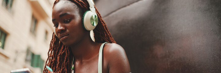 Gorgeous woman with African braids wearing top sits with headphones outside on the street and uses mobile phone, Panorama. Stylish girl listening to music on smartphone