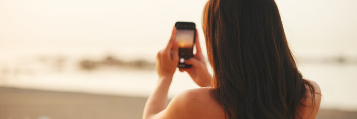Beautiful brown-haired woman with long hair is using mobile phone while standing on the beach,...