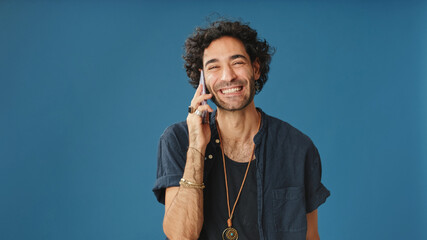Smiling man with curly hair, dressed in blue shirt, talking on mobile phone isolated on blue...