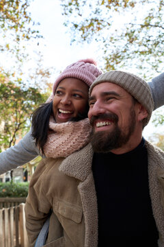 Vertical Portrait of beautiful multiracial happy couple outdoor young woman and man in love hugging and smiling in piggyback pointing somewhere at street in christmas time. People in winter clothes