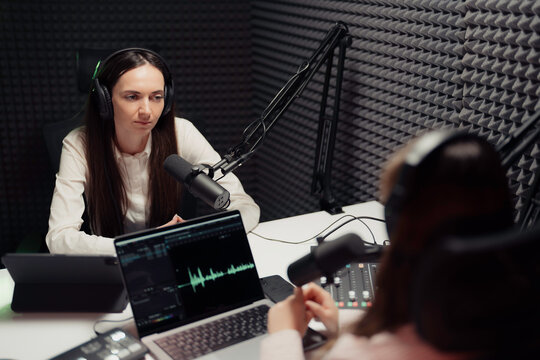 Two adult women engaged in podcast recording, one leading as a host, indoors, focused and confident, with positive energy, illustrating the concept of digital media production