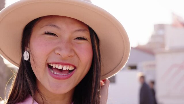 Close up of pretty young Asian generation z girl smiling joyful looking at camera. Chinese woman laughing posing cheerful standing outdoor on sunny summer vacation day. Beautiful happy female with hat