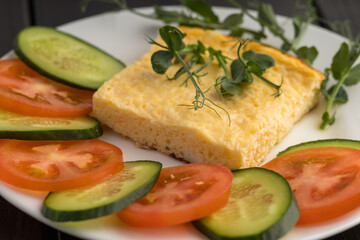 A white plate with a quiche slice, and fresh slices of tomato and cucumber, garnished with pea shoots