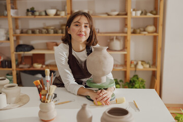Smiling entrepreneur crafts woman holding mug in pottery studio while looking at camera  - obrazy, fototapety, plakaty