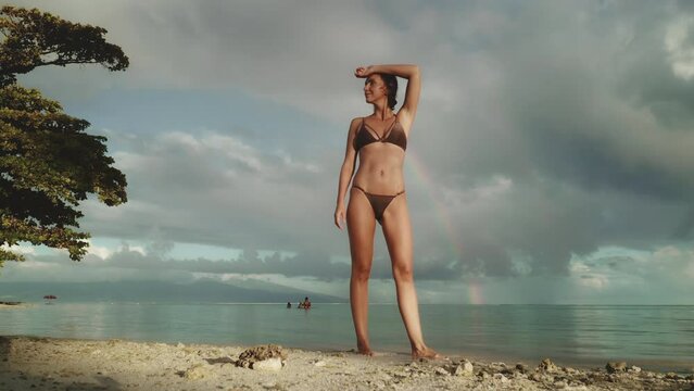 Young beautiful woman walking sunset sand beach. Sexy female in bikini enjoy tropical seascape under rainbow sky. Dramatic evening sunset after rain storm. Lifestyle travel, summer holiday vacation