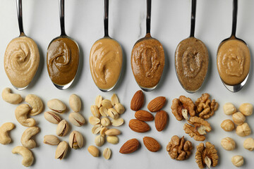 Tasty nut butters in spoons and raw nuts on white marble table, flat lay