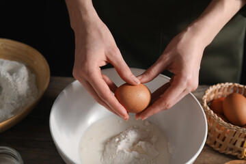 Making bread. Woman adding egg into dough at wooden table on dark background, closeup