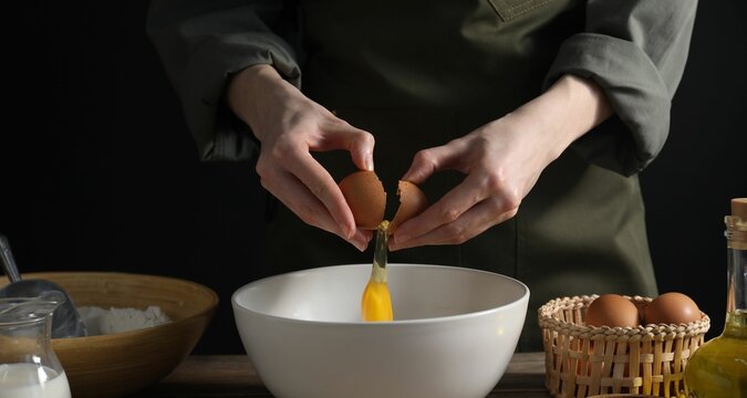 Making bread. Woman adding egg into dough at wooden table on dark background, closeup