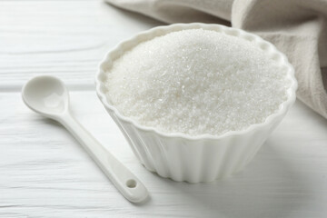 Fototapeta na wymiar Granulated sugar in bowl and spoon on white wooden table, closeup