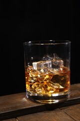 Whiskey with ice cubes in glass on wooden crate against black background, closeup