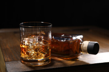 Whiskey with ice cubes in glass and bottle on wooden crate against black background, closeup