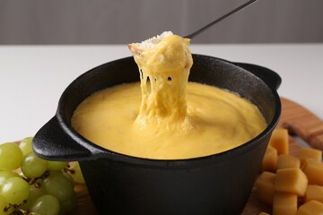 Dipping piece of bread into fondue pot with tasty melted cheese at table, closeup