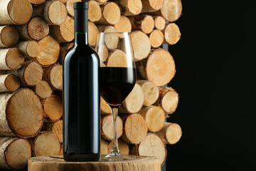 Stylish presentation of red wine in bottle and wineglass near wooden logs on black background,...