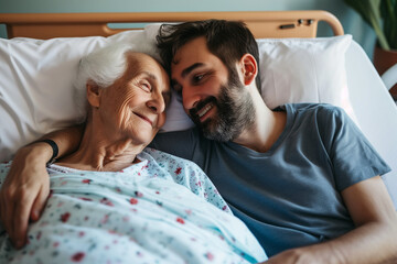 An elderly woman and a middle-aged man are embracing and smiling to each other in a hospital bed. - obrazy, fototapety, plakaty