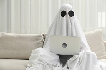 Creepy ghost. Person covered with white sheet using laptop on sofa at home