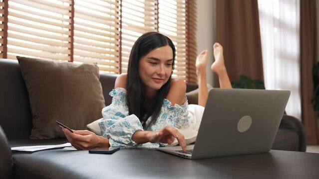 Young woman lying on sofa using laptop with credit card for internet banking, online shopping E commerce by online payment gateway at home. Modern and convenience online purchasing. Blithe