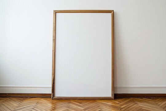 photo of a large blank frame leaning against a white wall. Frame is wood. resting on wooden parquet, close up, photo taken straight ahead