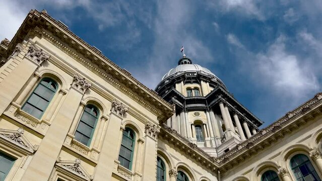 Rear views of the Illinois State Capitol Building in Springfield, Illinois, USA. Cloudy blue skies overhead. The US flag, State of Illinois Flag, and Black History Month Flag fly atop the dome.