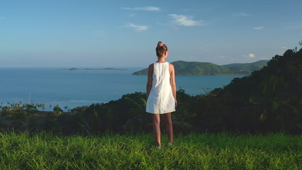 Woman enjoy fresh tropical island landscape standing on green grass hill looking on seascape horizon. Tourist female in white dress outdoor lifestyle travel on summer holiday vacation. Thailand, Samui