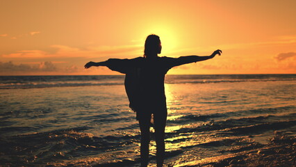 Woman raise hands up on sunset sea beach. Female silhouette stand on ocean coast, wind blow hair...