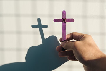 Male hand holding purple wooden cross with shadow. Christianity, faith, holy week and lent season concept.