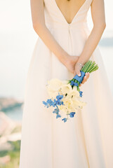 Bride stands with a bouquet of flowers in her hands folded behind her back. Cropped. Back view