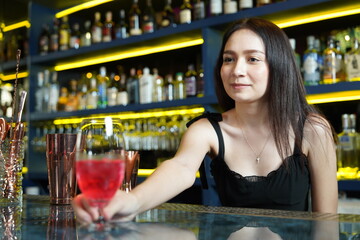Young professional Asian bartender works behind the bar counter delivering red wine glasses to customers in a nightclub.