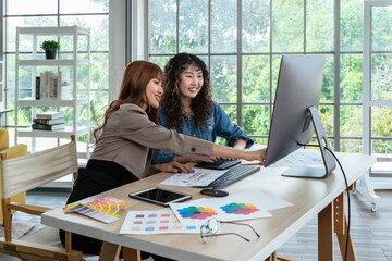 Two smiling young Asian female woman businessperson graphic designer working together, meeting and brainstorming, using computer in office, pointing at document.