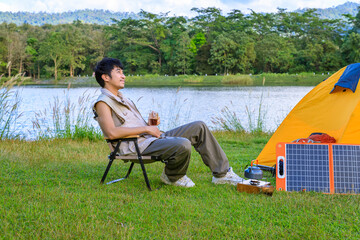 Asian male man sitting on chair, drinking coffee and feeling relax with camping tent beside lake and mountain, with mobile solar cell.