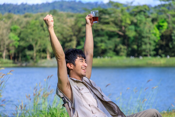 Asian male man sitting on chair, drinking coffee, feeling relax and raising hands, during camping beside lake and mountain.