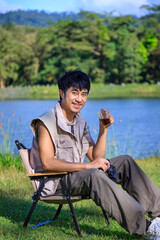 Young Asian male man sitting on chair, drinking coffee, feeling relax and raising hands, during camping beside lake and mountain. Looking at camera.
