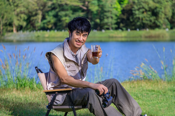 Young Asian male man sitting on chair, drinking coffee, feeling relax and raising hands, during camping beside lake and mountain. Looking at camera.