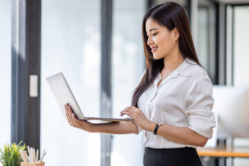 Portrait of Asian beautiful joyful woman holding laptop doing finance and standing in the office