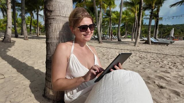Caucasian woman relaxing under palm trees using a digital tablet on a tropical beach resort during summer vacation