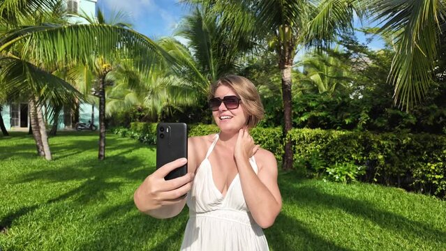 Happy woman in a white dress taking a selfie with a smartphone in a lush tropical garden on a sunny day, depicting a vacation or leisure concept