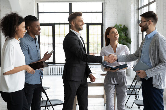 Group of young start-up founders meeting experienced business trainer at networking event in modern loft workspace. Diverse team of smiling office workers welcoming new colleague to company