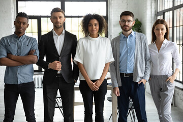 Group portrait of aspiring young coworkers. Confident start-up company employees at work standing together and looking at camera. Staff members of multinational organization in modern workspace - obrazy, fototapety, plakaty