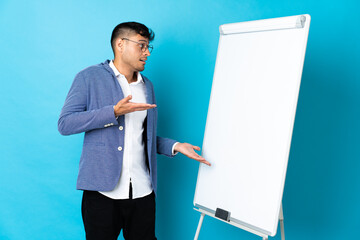 Young Colombian man isolated on blue background giving a presentation on white board and with...