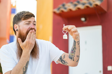 Redhead man with beard holding home keys at outdoors with surprise and shocked facial expression