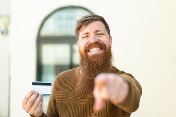 Redhead man with beard holding a credit card at outdoors points finger at you with a confident...