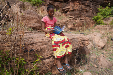 Young black elementary schoolgirl sitting on a rock reading in her textbook - Concept of female...