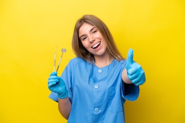 Dentist caucasian woman holding tools isolated on yellow background with thumbs up because something good has happened - obrazy, fototapety, plakaty